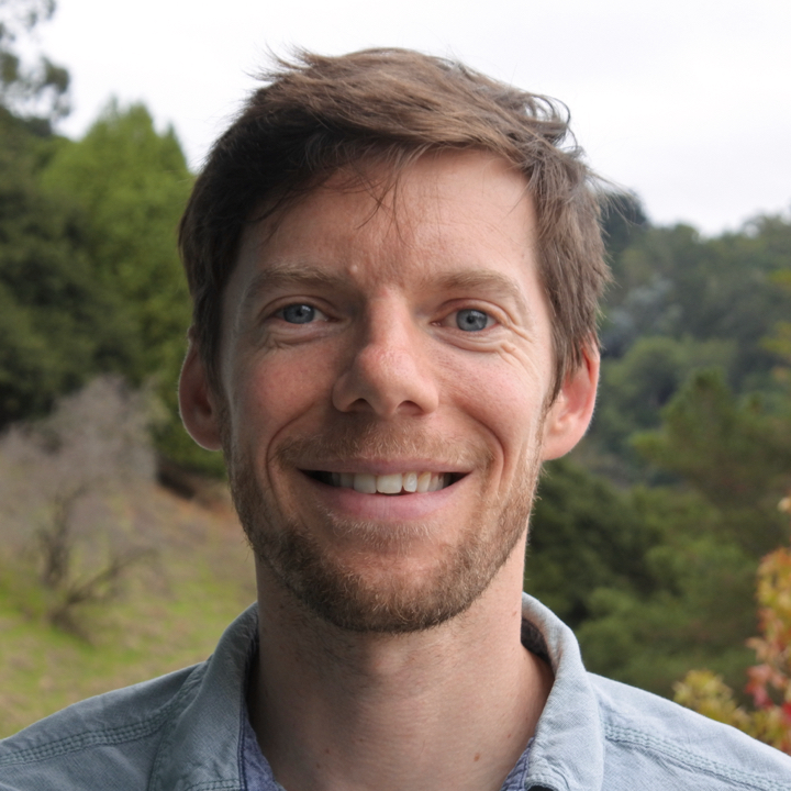 Trevor Keenan, a brown-haired person wearing a collared shirt, smiles for a headshot outdoors.