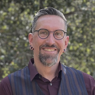 A person with short gray hair and a beard with glasses, wearing a dark purple collared shirt. Photographed outdoors.