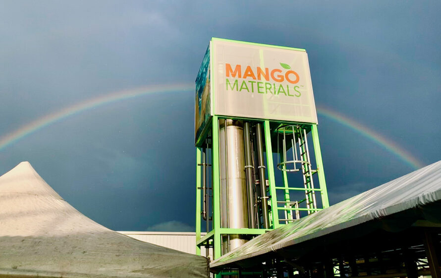 A chrome-colored methane storage tank featuring the Mango Materials logo rises above a rooftop, with a cloudy blue sky and rainbow in the background.