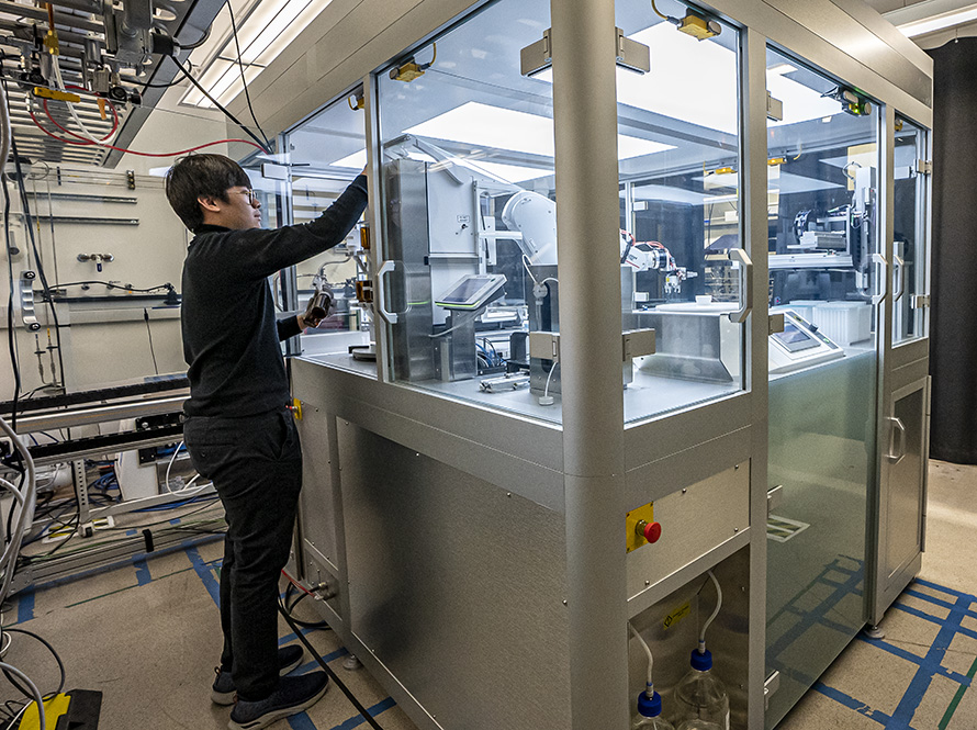 A dark-haired researcher stands at the left of a large, enclosed automated lab.