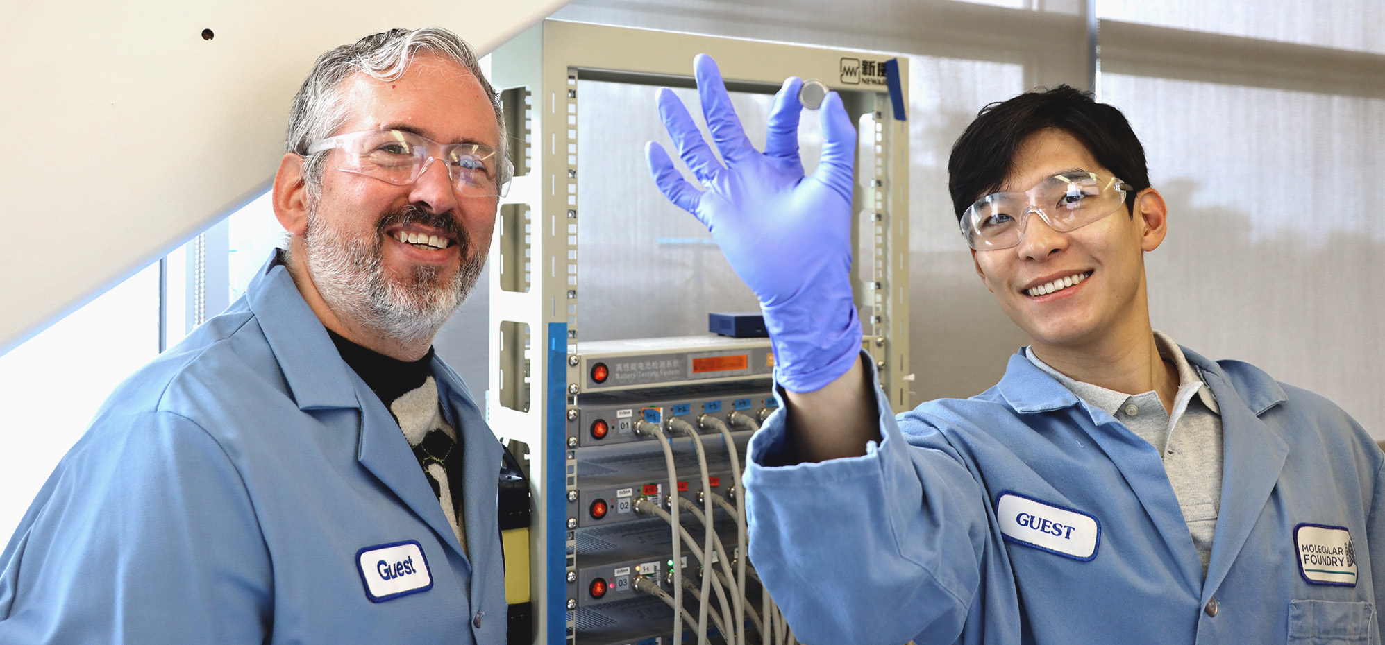 Two smiling people in blue lab coats and safety glasses. The person on the right has their arm raised in the center of the frame presenting a nickel-sized sample.
