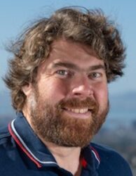 Alex Weber-Bargioni, a person with brown hair and beard wearing a navy collared shirt with white and red accents, smiling.