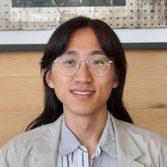 Portrait of Pinchen Xie, a person with long dark hair wearing glasses and a collared shirt and jacket, smiling.