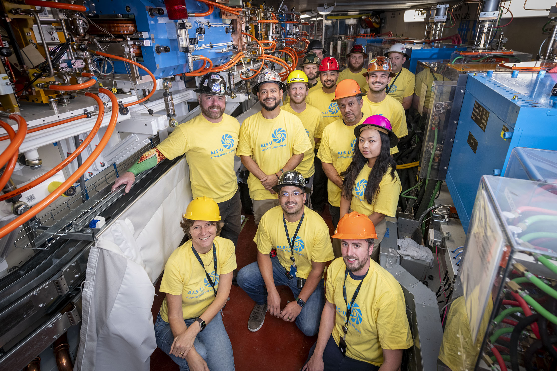 A group of people in yellow tshirts and multicolored hard hats. They are posing in a scientific facility.