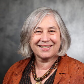 Marca Doeff, a person with bob-length silver hair wearing a burnt orange jacket over a black and red striped top and a matching necklace, smiling for a portrait.