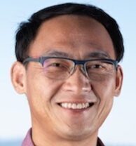 Yi Liu, a person with short black hair wearing glasses and a purple collared shirt, smiling.