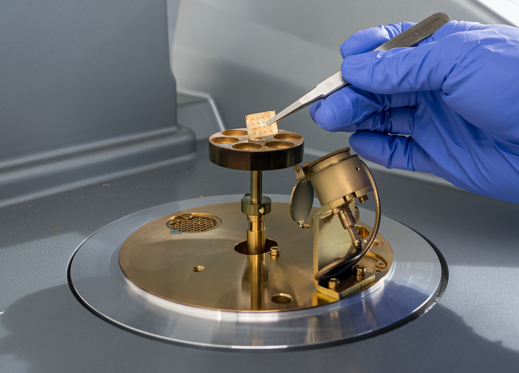 A gloved hand uses tweezers to place a small sample into a scientific instrument with metallic components.