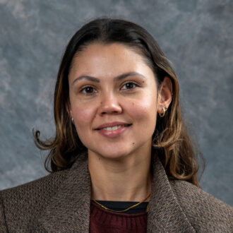 Julia Correa, a person with long brown hair wearing a brown blazer over a maroon sweater, smiling for a portrait.