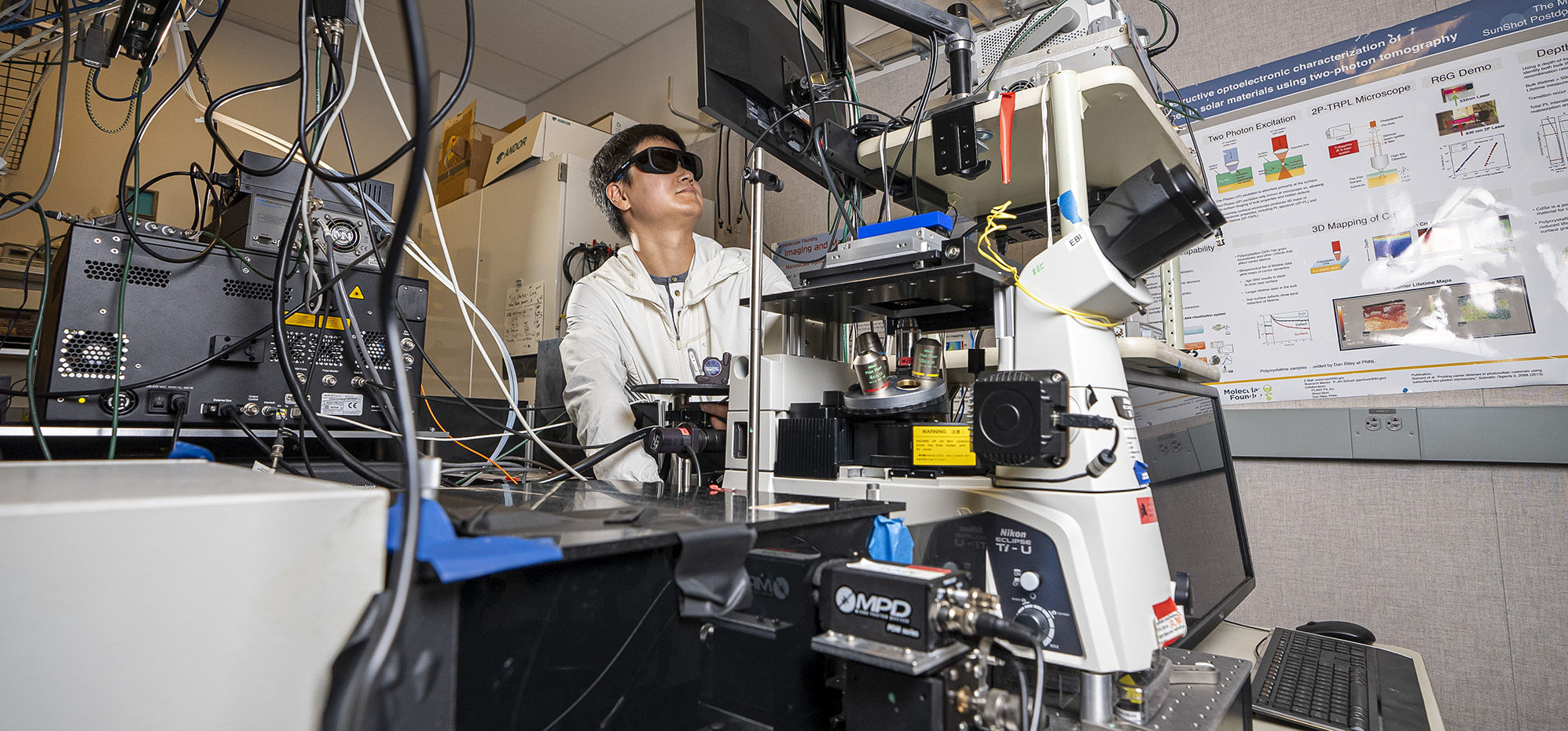 Scientist in protective eyewear adjusts a microscope in a high-tech lab filled with optical and electronic equipment.